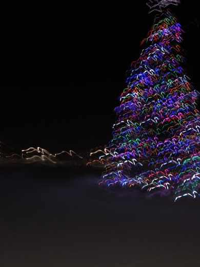 Long exposure photos of a lit Christmas tree in the night, leaving a colorful trail of vibrant, zig-zagging, lights in front of a black background