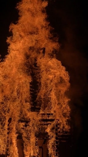 A large wooden structure burns up in a bonfire, the number "21" visible amidst the flurry of embers, flames, and ash